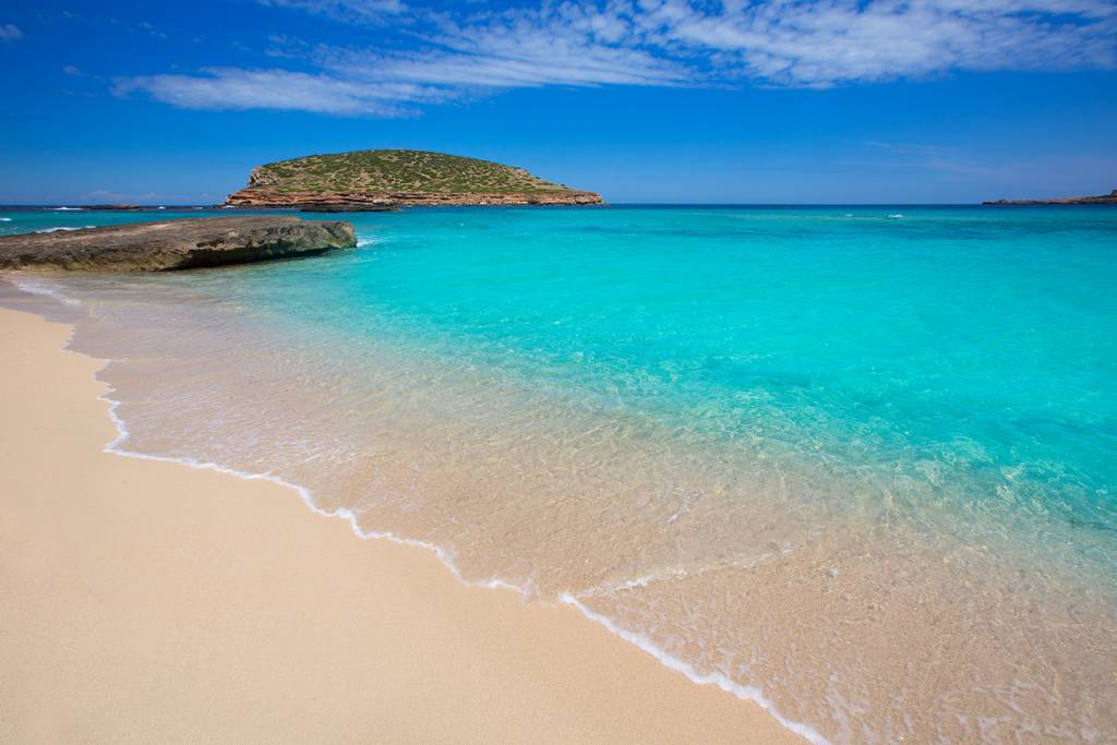 Spiaggia paradisiaca di Cala Comte dalle acque turchesi e cielo limpido, con un isolotto roccioso sullo sfondo e dolci onde che si infrangono sulla riva