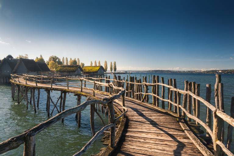 Ein rustikaler Holzsteg führt über einen ruhigen See zu charmanten Hütten mit Reetdächern unter einem klaren blauen Himmel.
