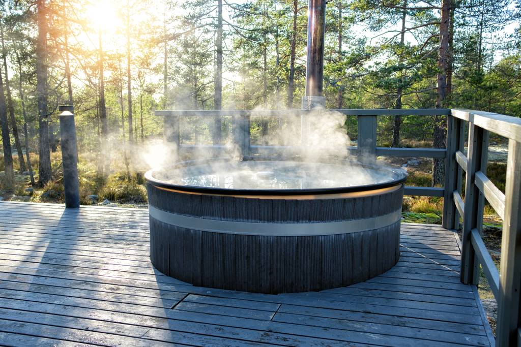 A steaming hot tub on a wooden deck in a forest setting, with sunlight filtering through the trees.