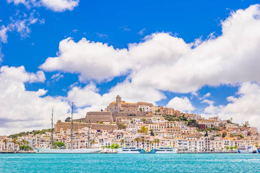 Paisaje costero de una ciudad con una fortaleza en lo alto de la colina, bajo un cielo azul con nubes esponjosas, frente a aguas turquesa y con barcos en el puerto.