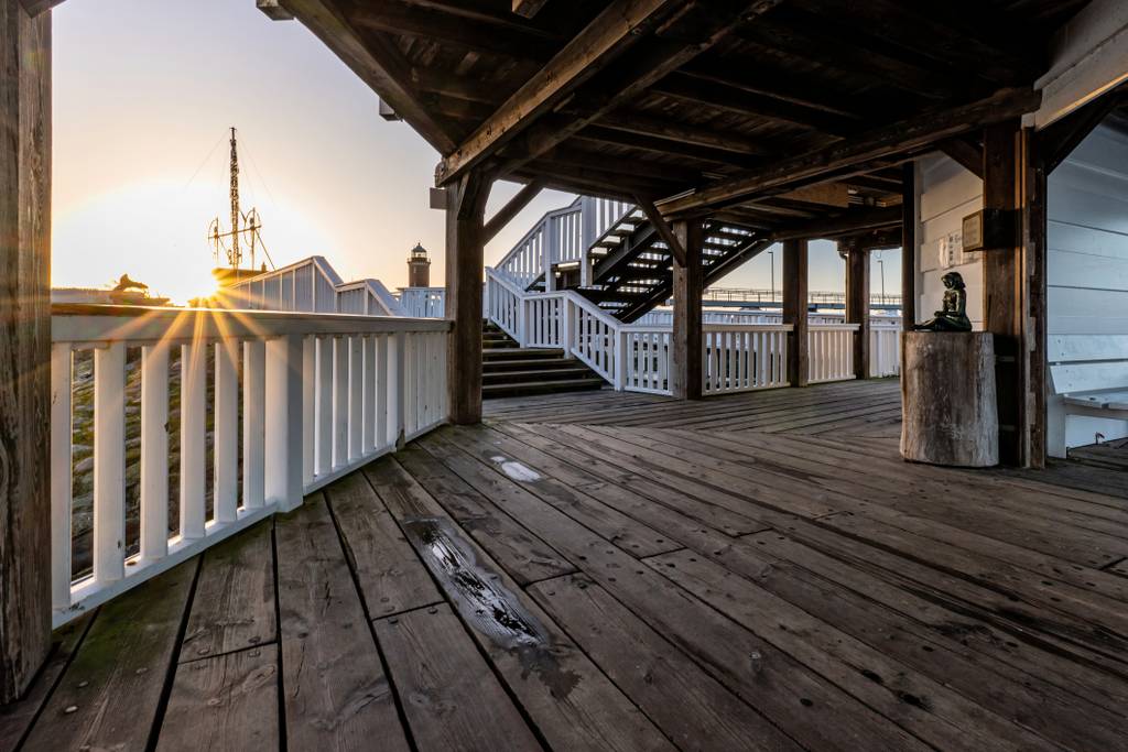 Sonnenuntergang von einem verwitterten hölzernen Pierdeck aus, unter einem überdachten Steg mit weißen Geländern, Treppen und einem fernen Schiffsmast.