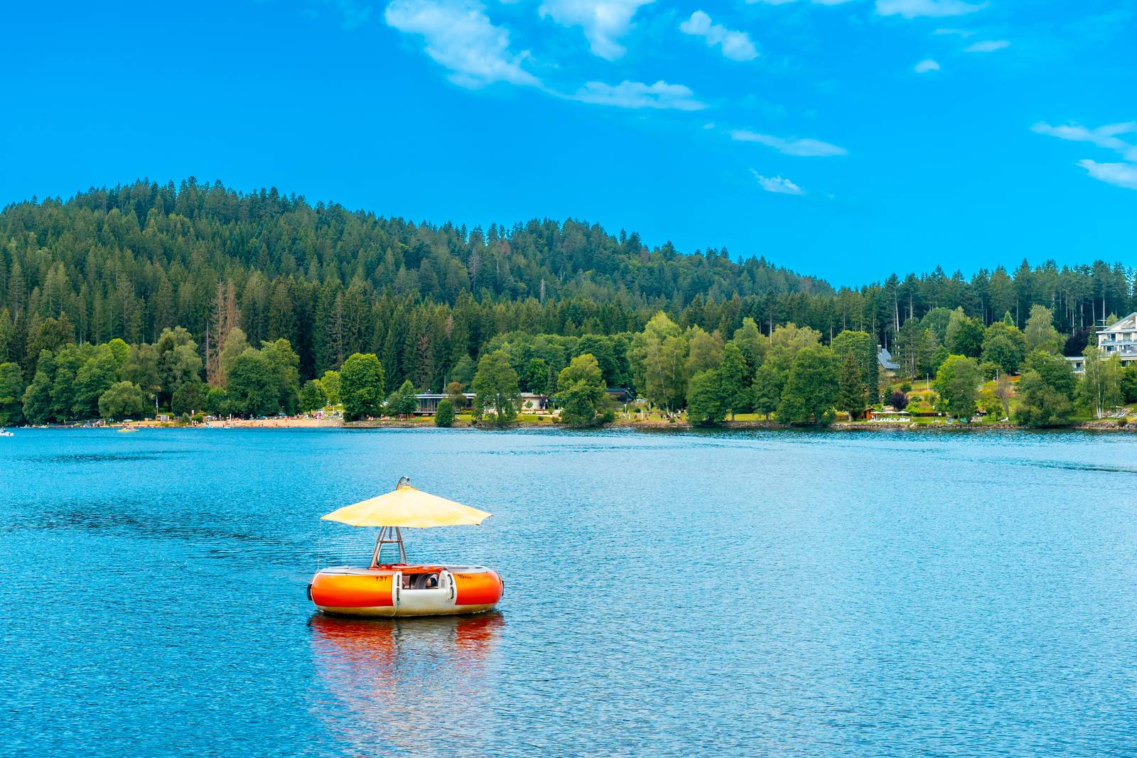 Ein kleines Floß mit einem Sonnenschirm treibt auf einem ruhigen See, umgeben von dichtem, grünem Wald unter klarem blauem Himmel.