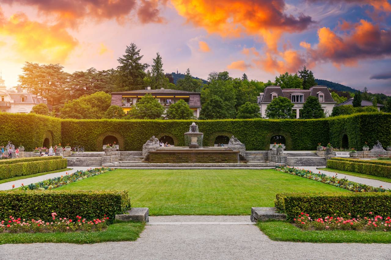 Eleganter Garten mit sorgfältig geschnittenen Hecken, einem zentralen Springbrunnen und umliegenden historischen Gebäuden, vor einem farbintensiven Sonnenuntergangshimmel.