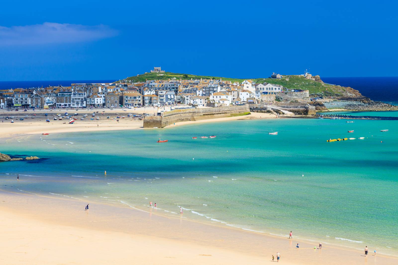 A coastal town with sandy beaches, turquoise water, and scattered boats under a clear blue sky. White buildings sit on a green hill in the background.