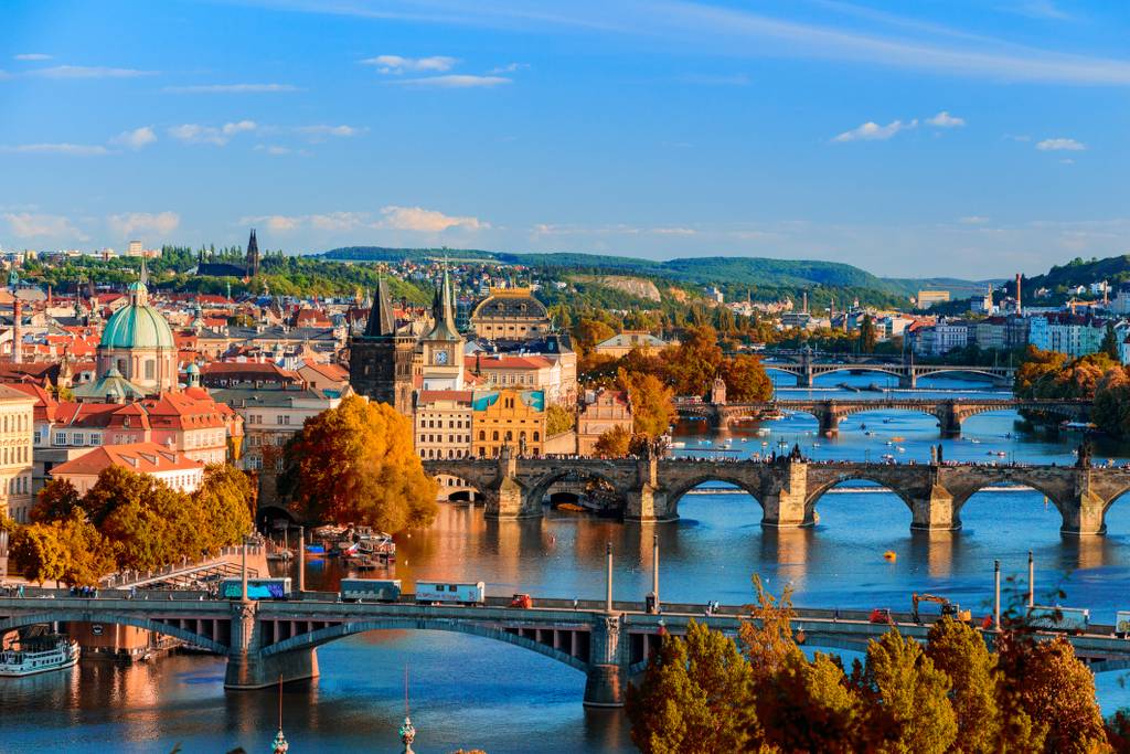 Aerial view of Prague with colorful autumn trees, multiple bridges crossing the Vltava River, and historic buildings under a clear blue sky.