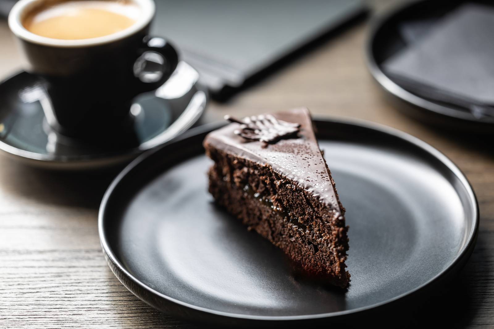 A slice of Sacher Torte on a black plate next to a cup of coffee on a wooden table.