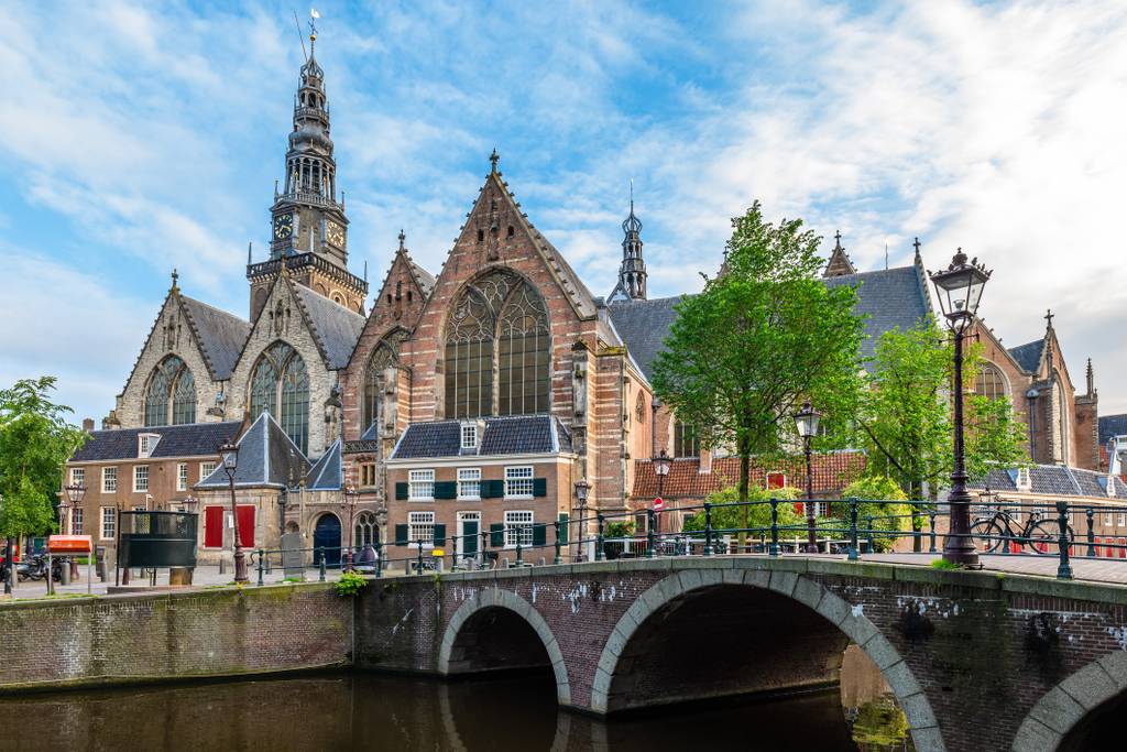 Historic Gothic church with a large central stained glass window, tall spire, and surrounding buildings, viewed from a nearby canal and bridge.