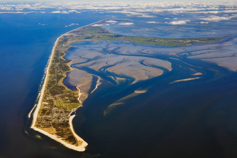 Luftaufnahme eines schmalen, sandigen Küstenstreifens, der ins Meer hineinragt, umgeben von blauem Wasser und sandigen Wattflächen, mit vereinzelten Wolken am Himmel.