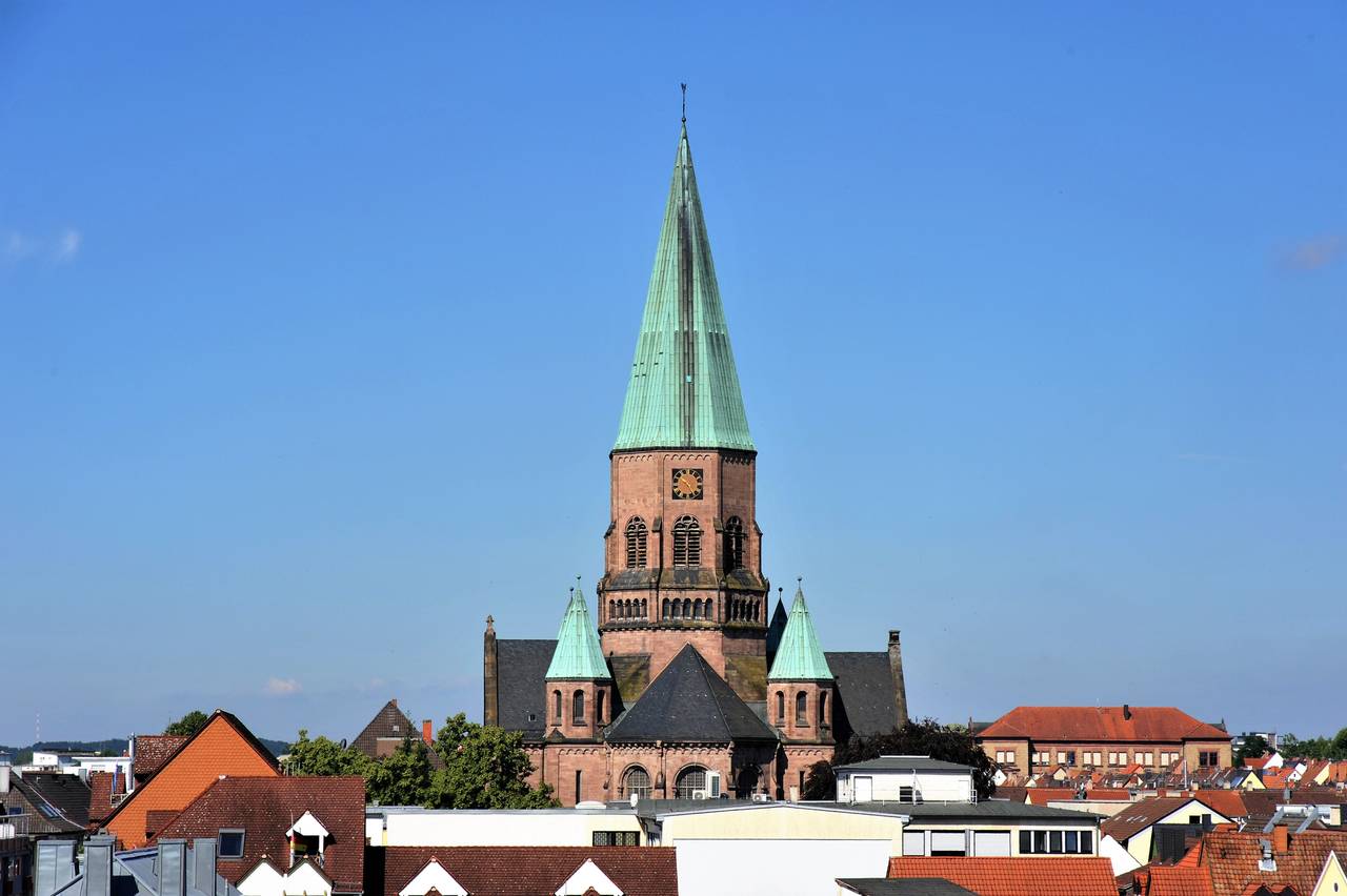 Eine historische Kirche mit einem hohen, grünen Spitzturm und zwei kleineren Türmen vor klarem blauen Himmel, umgeben von Hausdächern.