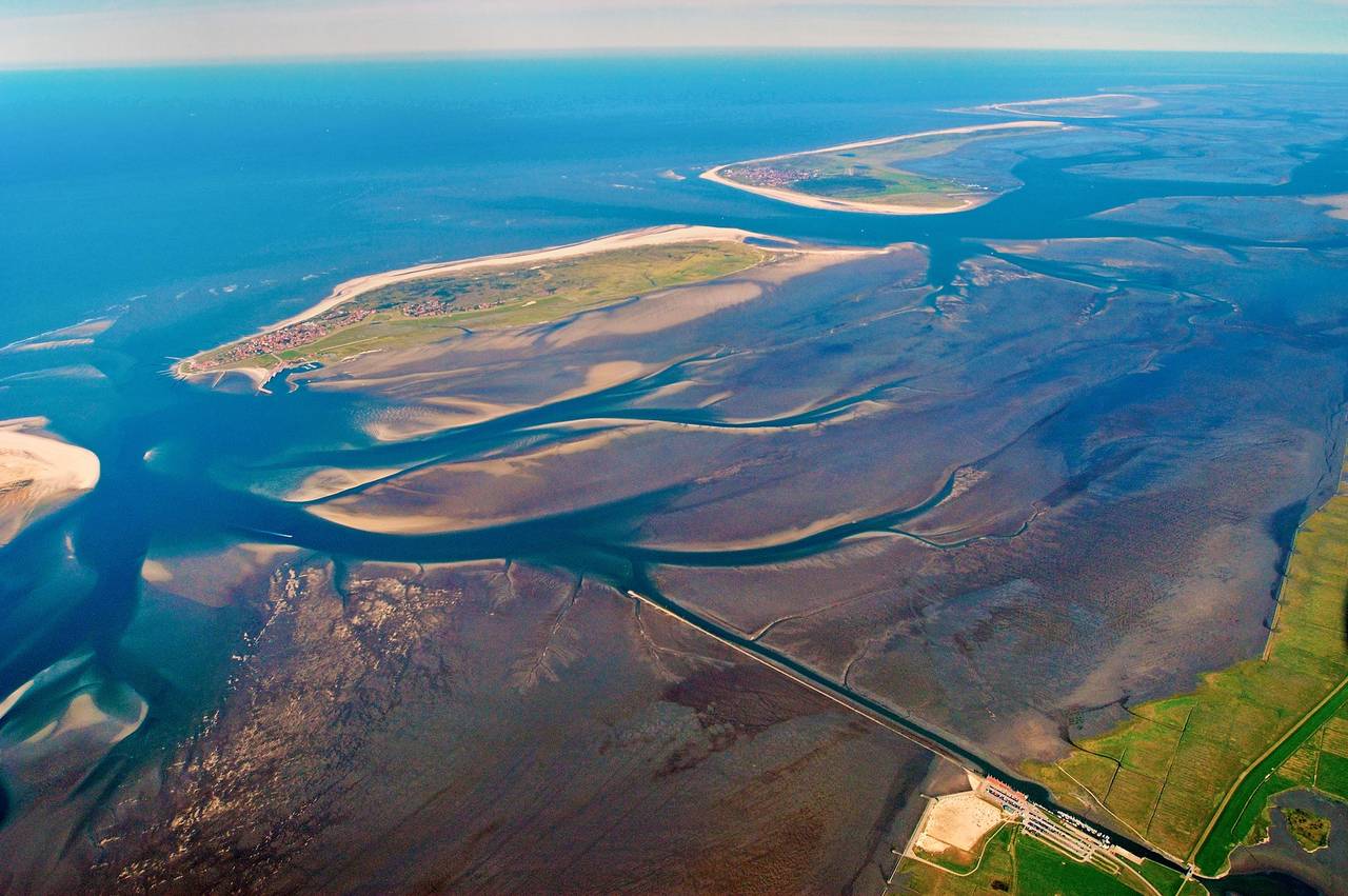 Luftaufnahme von Wattflächen und Inseln mit Sandbänken und Prielen, die ins Meer übergehen, umgeben von einer entfernten Küstenlinie und grünen Landflächen.