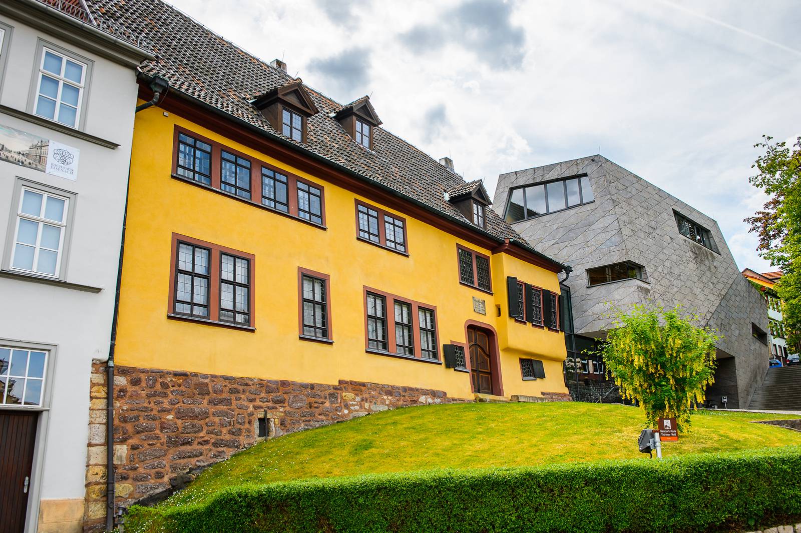 Historisches gelbes Gebäude mit dunklen Fenstern neben einem modernen grauen Bauwerk, gelegen an einem grasbewachsenen Hang unter einem bewölkten Himmel