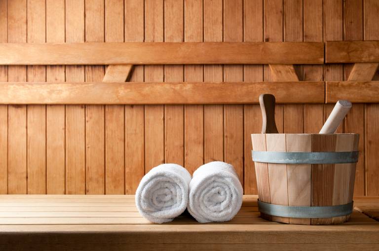 Ein Innenraum einer Holzsauna mit zusammengerollten weißen Handtüchern sowie einem Holzeimer mit Kellen, die auf einer Bank platziert sind.