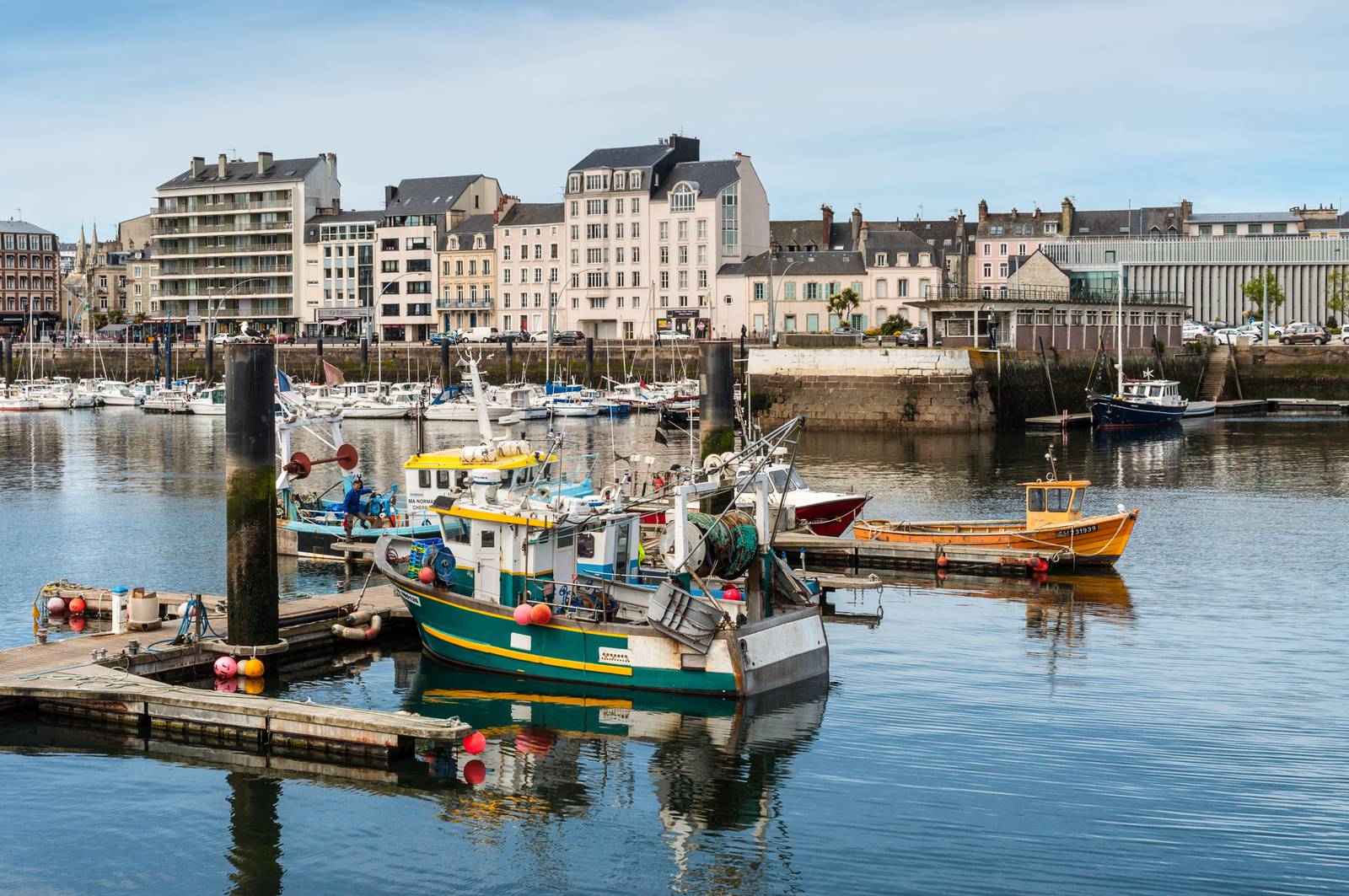 Fishing boats docked in a calm harbor, with a backdrop of multi-story buildings and a clear sky.
