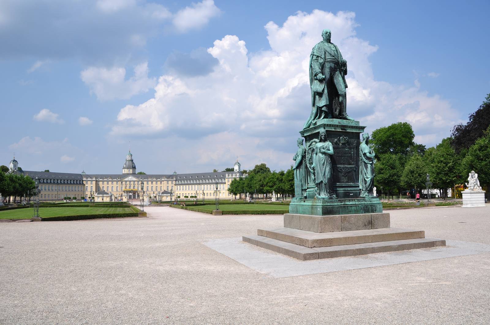 Bronzestatue auf einem Sockel in einem Park mit einem prächtigen Gebäude und wolkigem blauen Himmel im Hintergrund