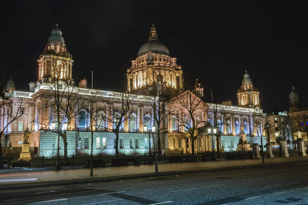 Illuminated historic building with domes and columns at night, trees silhouetted in the foreground, and a dark sky above.