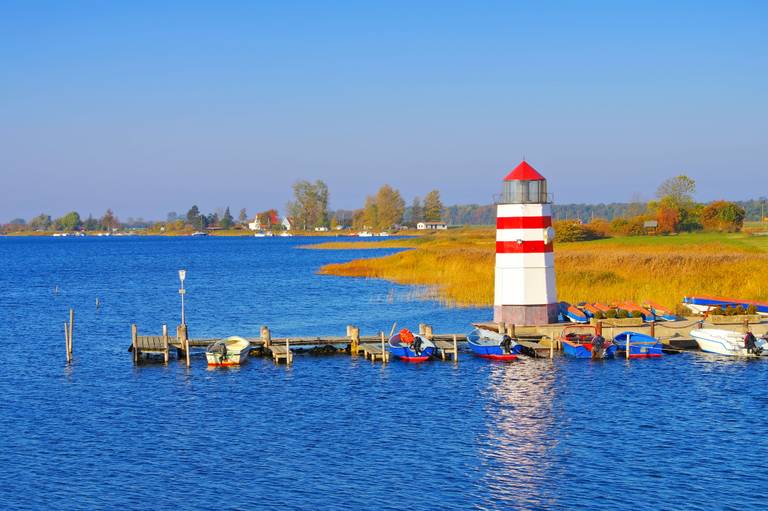 Leuchtturm mit roten und weißen Streifen neben einem festgemachten Ruderboot an einem See, eingerahmt von herbstlichem Laub und einem klaren blauen Himmel.