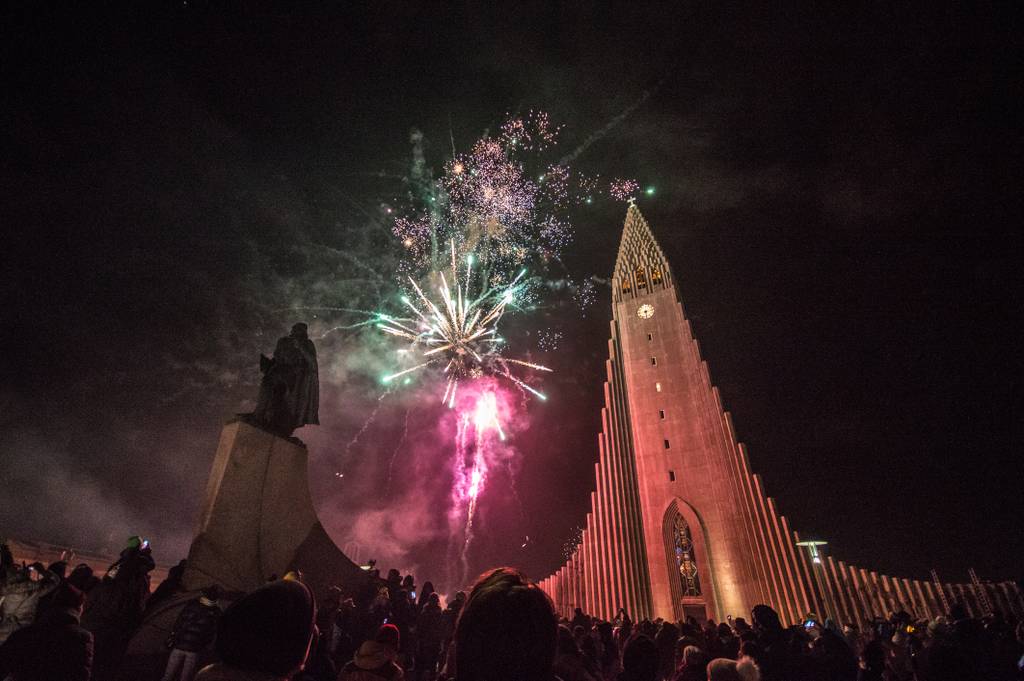 Fireworks illuminate the night sky above a large, illuminated church with a pointy roof, surrounded by a crowd and a statue in the foreground.