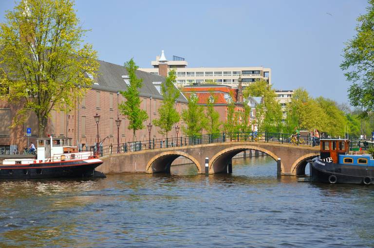 Ein malerischer Blick auf einen Kanal in Amsterdam mit einem Bogenbrücke, Booten, historischen Gebäuden und grünen Bäumen unter klarem blauem Himmel.