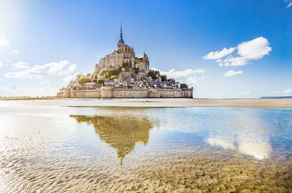 Une abbaye insulaire, avec sa flèche imposante et son architecture médiévale, se reflétant dans des eaux calmes et peu profondes, sous un ciel d'un bleu éclatant parsemé de quelques nuages.