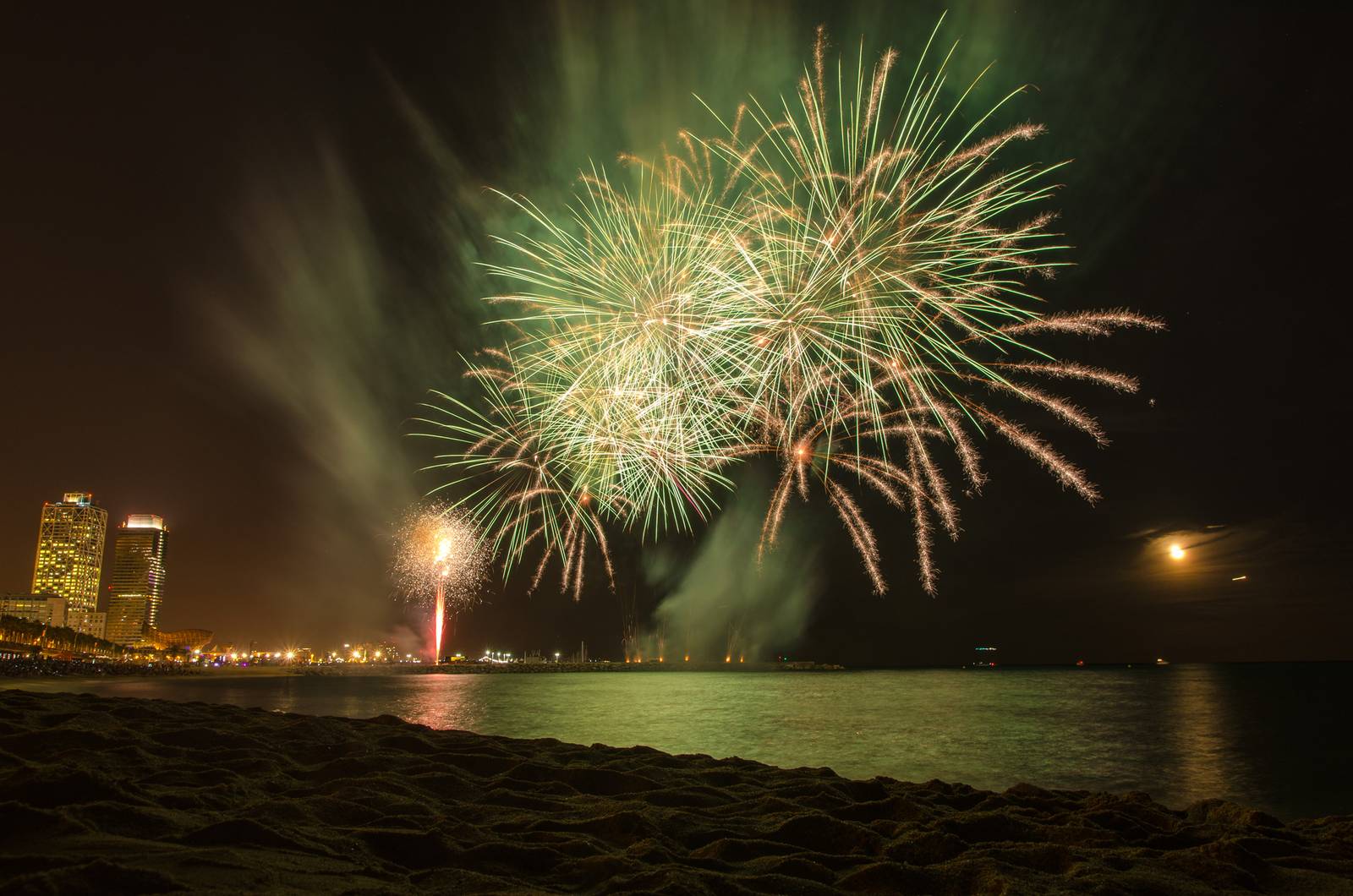 Feuerwerk über einem Stadtstrand bei Nacht, grüne und weiße Explosionen erleuchten den Himmel und spiegeln sich im Wasser.