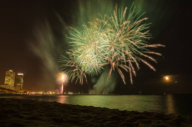 Feuerwerk über einem Stadtstrand bei Nacht, grüne und weiße Explosionen erleuchten den Himmel und spiegeln sich im Wasser.