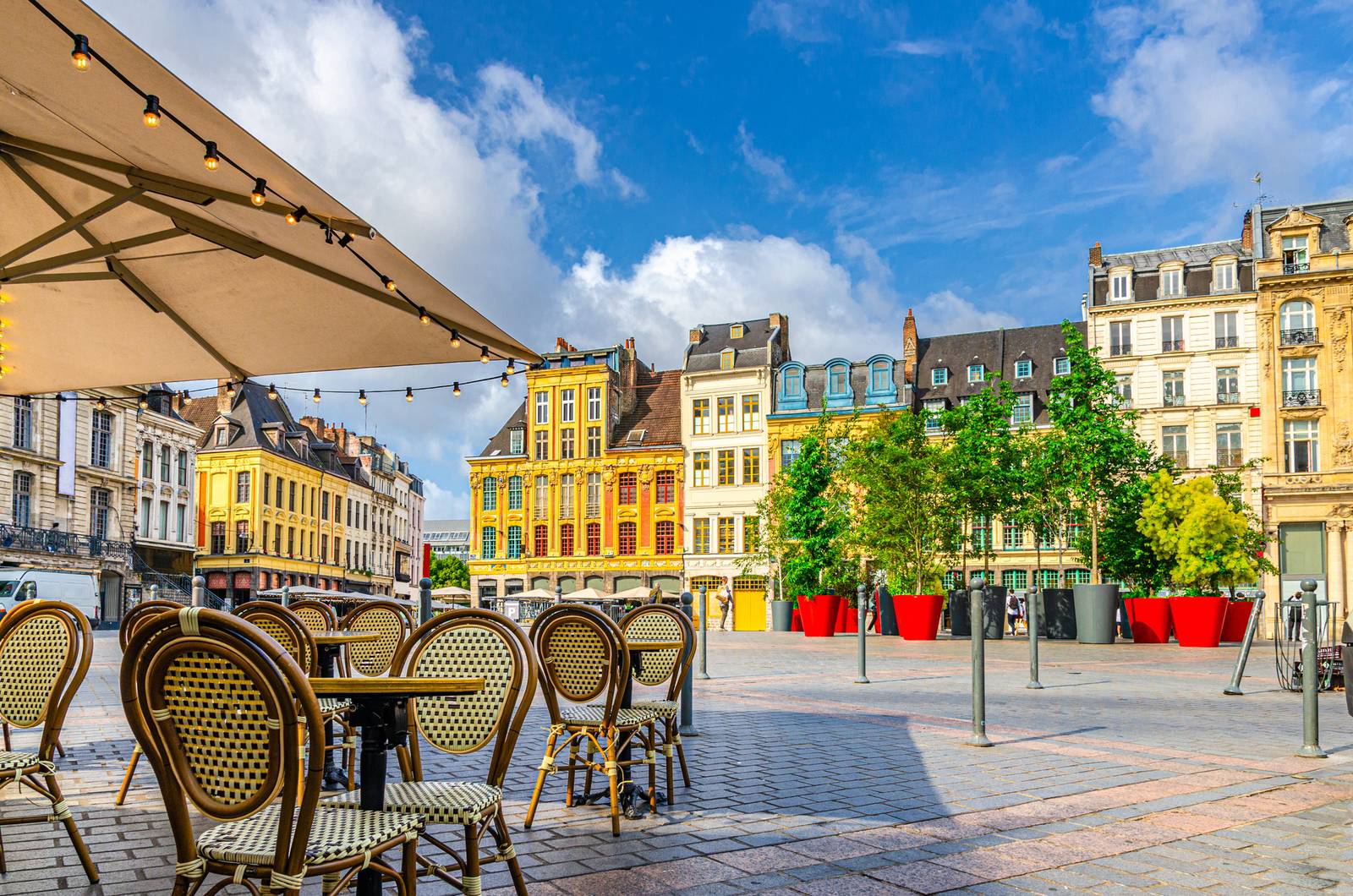 Outdoor café with wicker chairs under an umbrella on a cobblestone plaza, surrounded by colorful historic buildings and large potted trees.