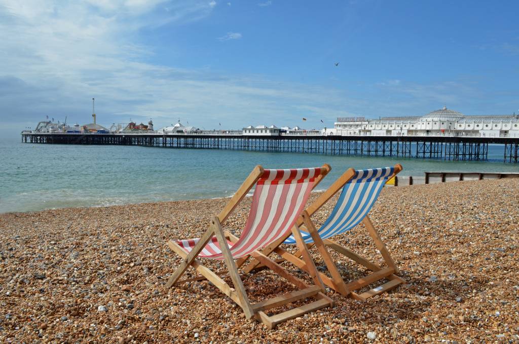 Two striped deck chairs on a pebbly beach face the sea, with a long pier extending into the water under a partly cloudy sky.