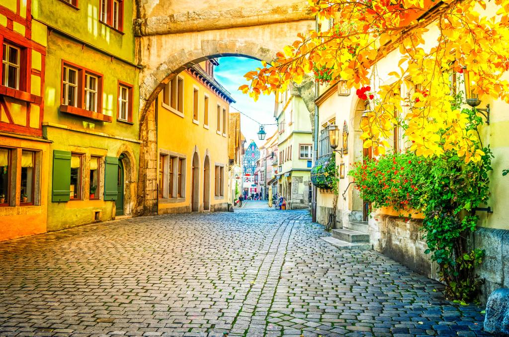 Eine malerische Kopfsteinpflasterstraße mit bunten historischen Gebäuden, einem Torbogen und leuchtenden Herbstblättern unter einem blauen Himmel.