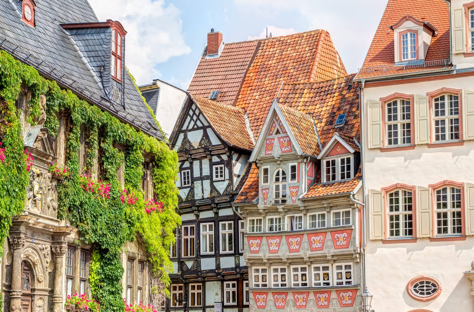 Bunte, historische europäische Gebäude mit kunstvoll verzierten Fassaden und roten Ziegeldächern, geschmückt mit Grünpflanzen und Blumen, unter einem teilweise bewölkten Himmel