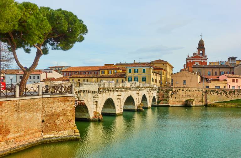 Historische Steinbrücke über einem ruhigen Fluss, mit bunten Gebäuden und einem Kirchturm im Hintergrund, unter klarem Himmel
