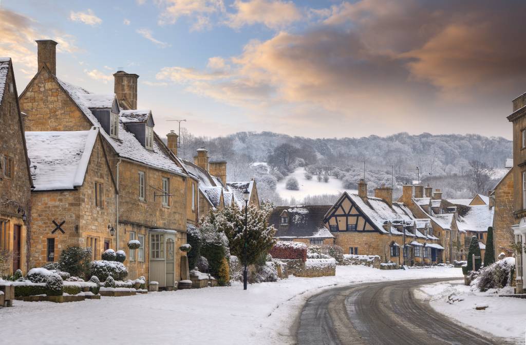 Snow-covered village street lined with stone cottages, under a colorful sky with hills in the background.