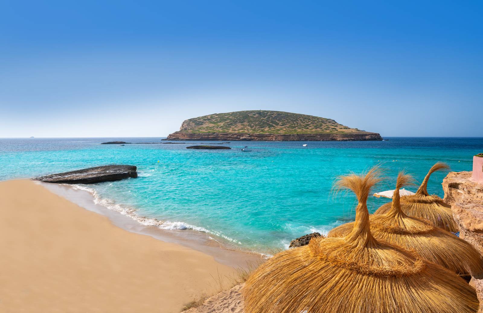 Sandstrand mit Strohmsonnenschirmen, Blick auf türkisfarbenes Wasser und eine kleine Insel unter klarem, blauem Himmel