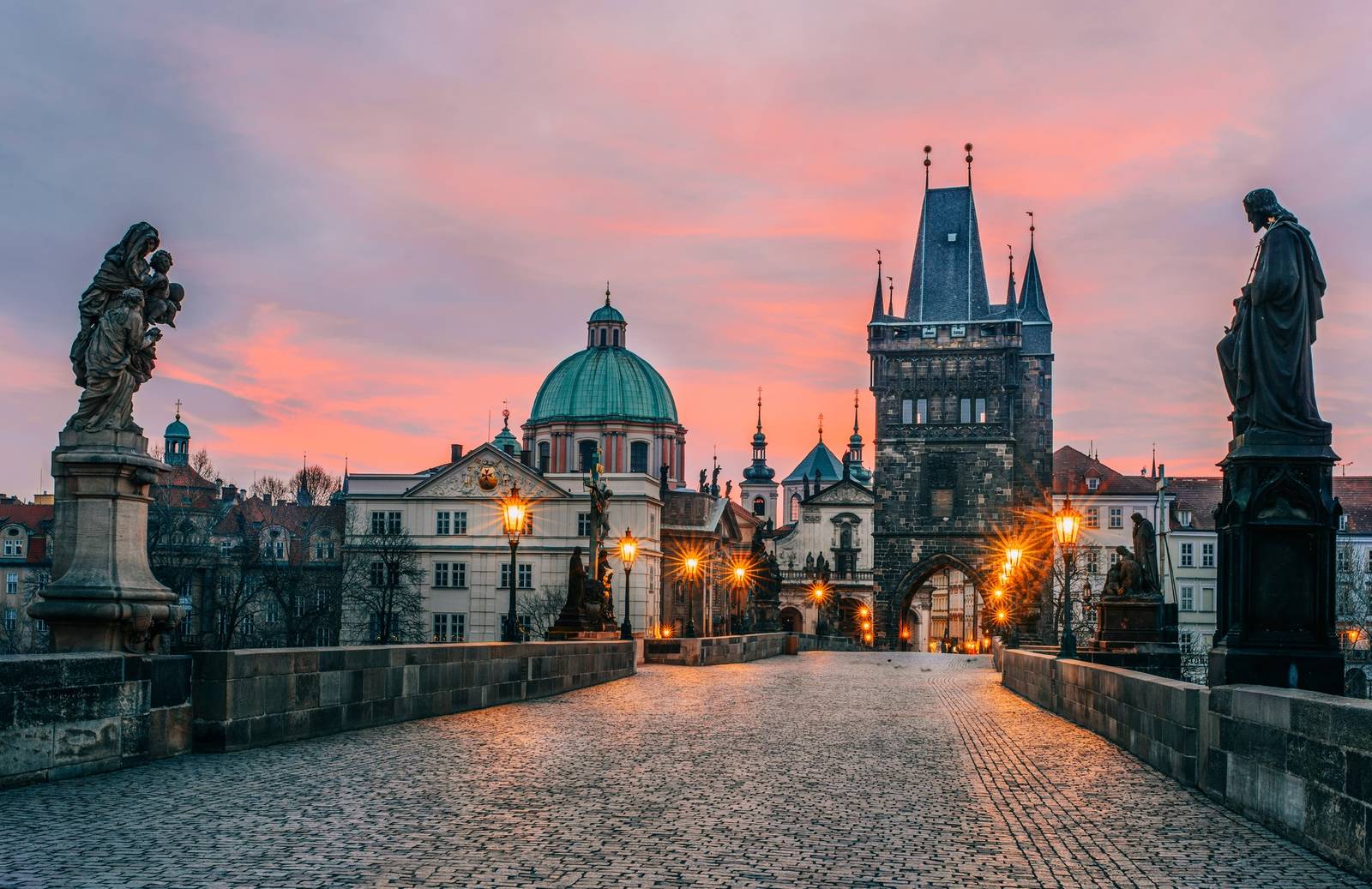 Die Karlsbrücke im Morgengrauen mit silhouettierten Statuen, Kopfsteinpflaster und beleuchteten Türmen der Altstadt unter einem rosa-blauen Himmel.