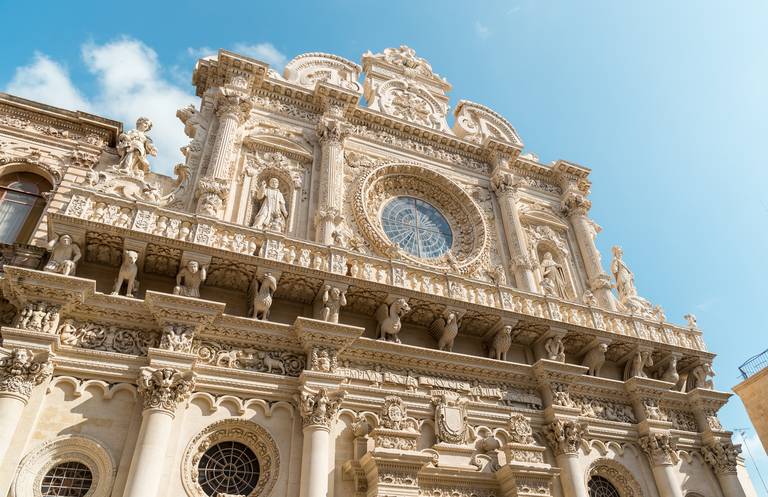 Verschnörkelte Barockfassade mit kunstvollen Schnitzereien, Statuen und einer großen Fensterrose unter strahlend blauem Himmel.