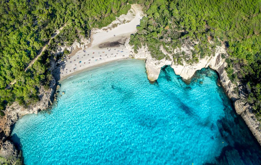 Vista aerea di  Cala Mitjana, una spiaggia isolata dalle acque turchesi, circondata da vegetazione lussureggiante e scogliere, con lettini sparsi sulla sabbia.