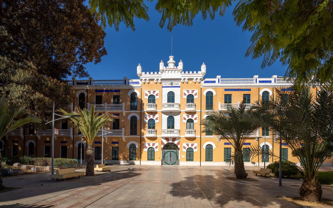 Edificio histórico de color amarillo con detalles en blanco bajo un cielo azul claro, rodeado de palmeras y una plaza pavimentada con bancos.