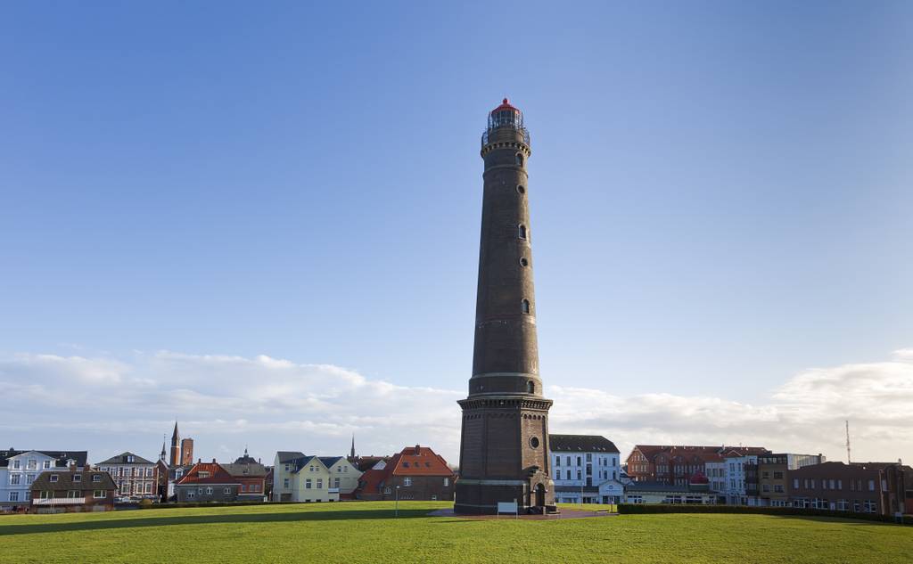 Hoher Leuchtturm aus Ziegelstein mit roter Laterne auf einer grünen Rasenfläche, dazu eine Reihe farbenfroher Küstenhäuser und ein klarer blauer Himmel.