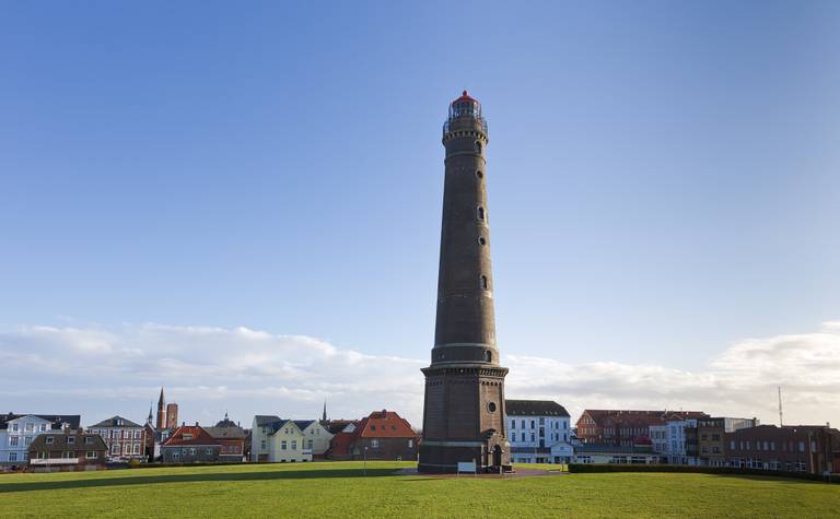 Hoher Leuchtturm aus Ziegelstein mit roter Laterne auf einer grünen Rasenfläche, dazu eine Reihe farbenfroher Küstenhäuser und ein klarer blauer Himmel.