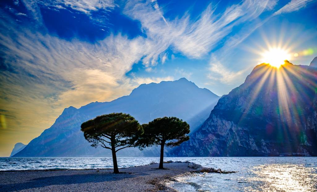 Zwei Bäume an einem Strand, dahinter erheben sich Berge. Am blauen Himmel ziehen feine, zarte Wolken vorbei, während die Sonne über den Berggipfeln hervorscheint.