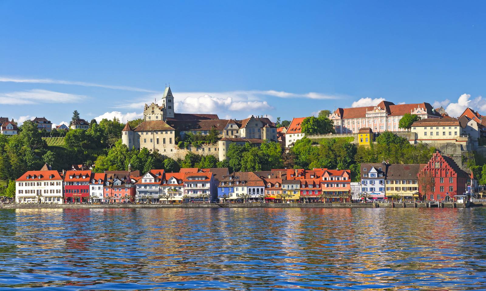 Bunte Häuser säumen das Ufer unter klarem, blauem Himmel, im Hintergrund ein historisches Kirchgebäude und üppiges Grün auf einem Hügel