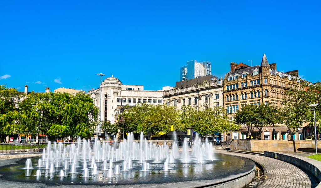 Fountain with jets of water surrounded by historical buildings in Manchester and trees under a clear blue sky.