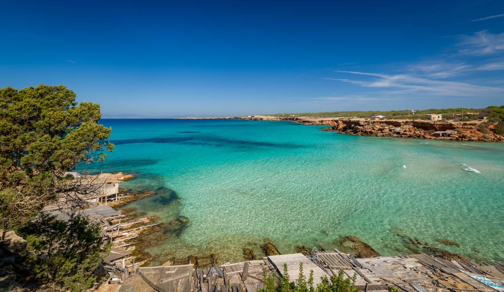 Cala aona, Baia dalle acque turchesi con casette in legno sulla spiaggia, costa rocosa e pini sotto un cielo azzurro limpido.
