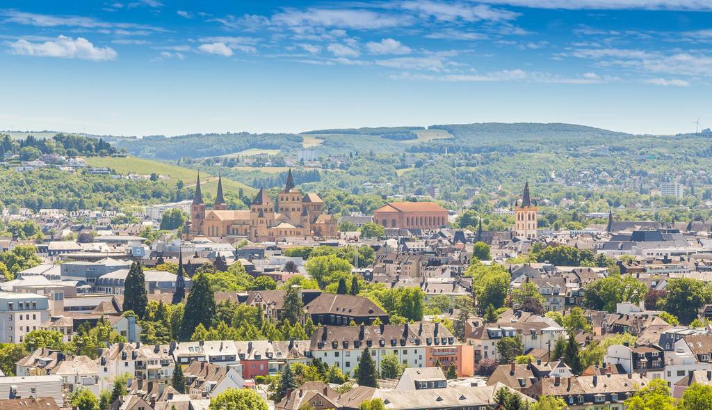 Malerischer Blick auf eine Stadt mit historischen Gebäuden, Türmen und üppigem Grün, eingebettet in sanfte Hügel unter einem blauen Himmel mit vereinzelten Wolken.