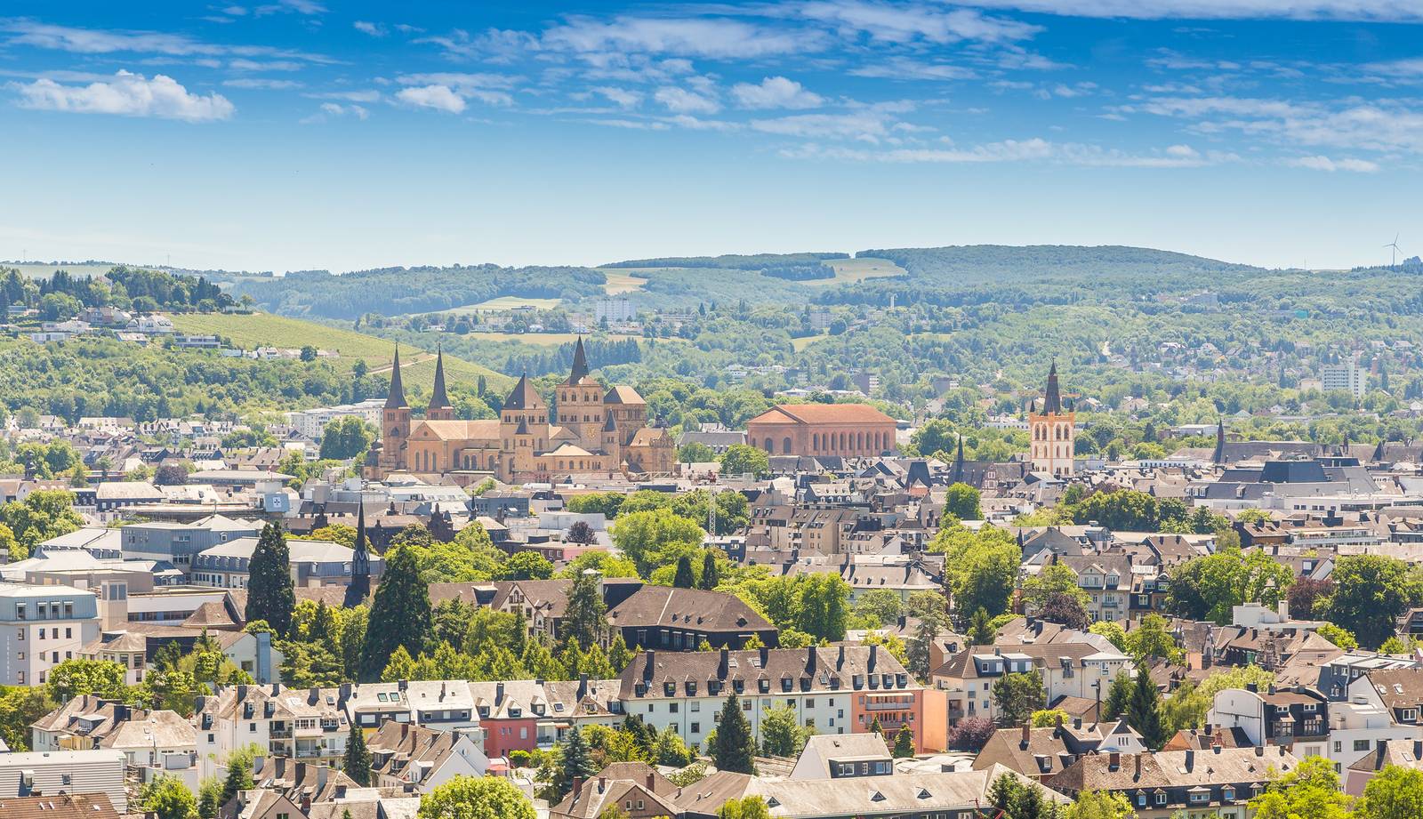 Luftaufnahme einer Stadtlandschaft mit historischen Gebäuden, üppigem Grün und fernen, sanften Hügeln unter strahlend blauem Himmel