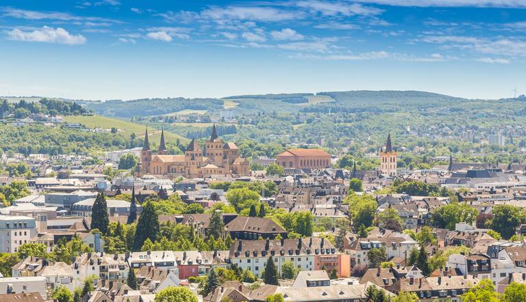 Luftaufnahme einer Stadtlandschaft mit historischen Gebäuden, üppigem Grün und fernen, sanften Hügeln unter strahlend blauem Himmel