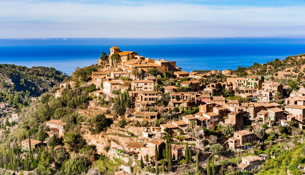 Borgo di Deià, in cima a una collina con vista sul mare, circondato dalla vegetazione e da case con tetti in terracotta sotto un cielo azzurro limpido.