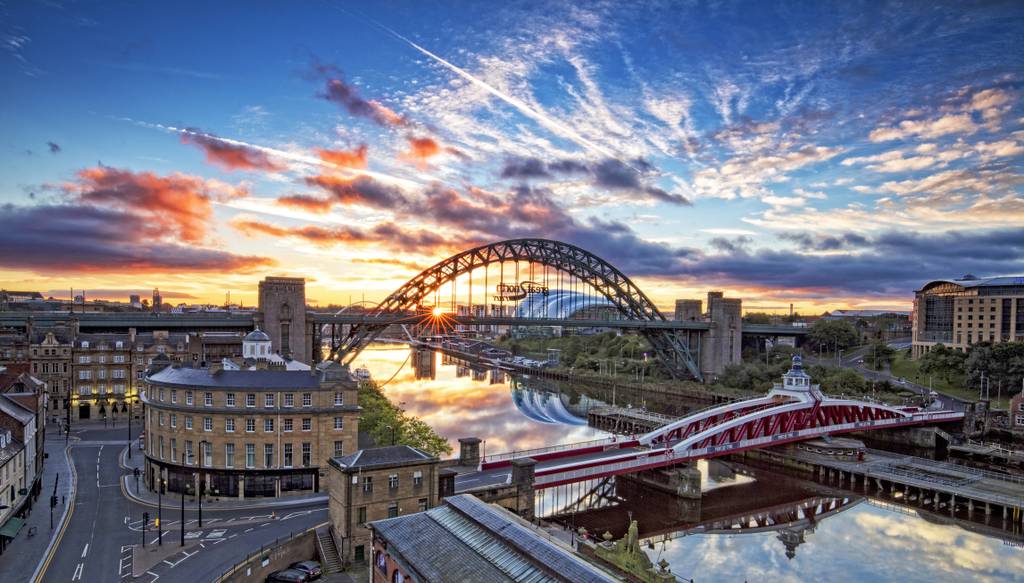 Sunset view of the Tyne Bridge spanning a river, with vivid clouds and reflections in the water, surrounded by historic buildings and a red bridge.