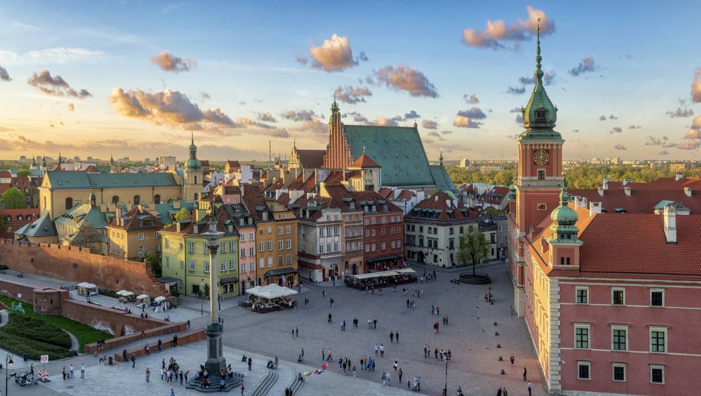 Aerial view of a historic square in a city with colorful buildings, a tall clock tower, and scattered clouds in a blue sky at sunset.