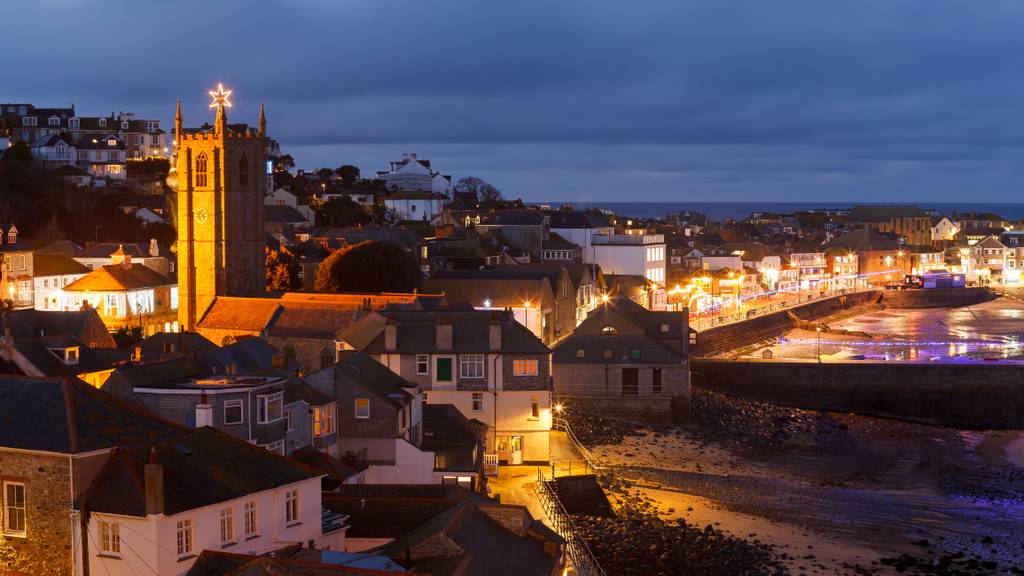A scenic coastal town at dusk, with illuminated buildings, a prominent church tower, and reflections on the water under a cloudy sky.