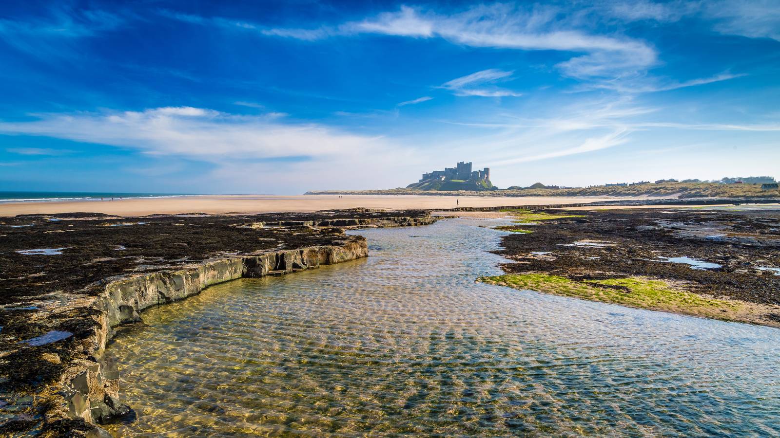 A scenic view of a rocky shoreline with a clear tidal pool, distant beach, and a castle under a blue sky with wispy clouds.